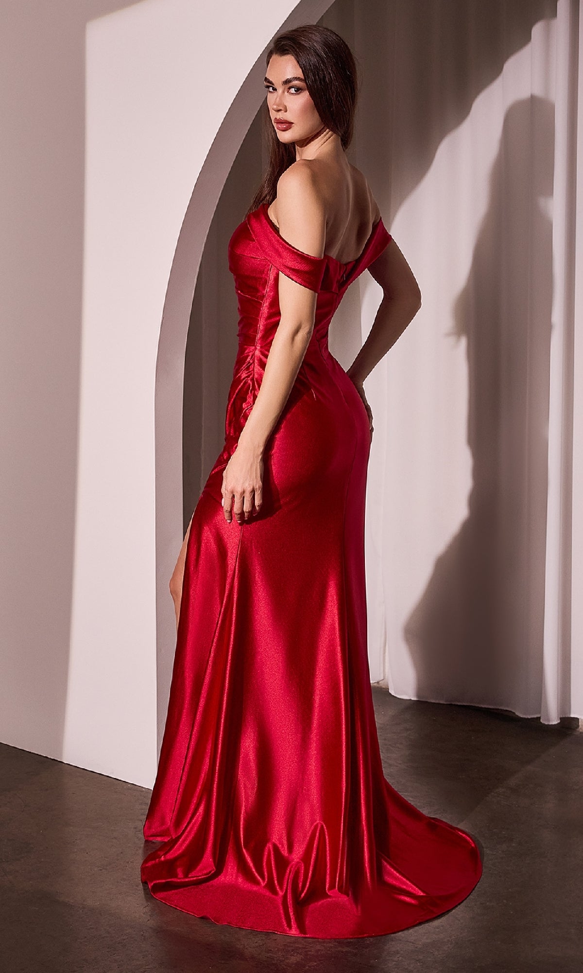 A woman wearing the Ladivine J877 Off-the-Shoulder Long Prom Dress in shiny red with a mermaid skirt stands sideways, gazing over her shoulder, in a minimalist room with white drapes and an arched wall feature.